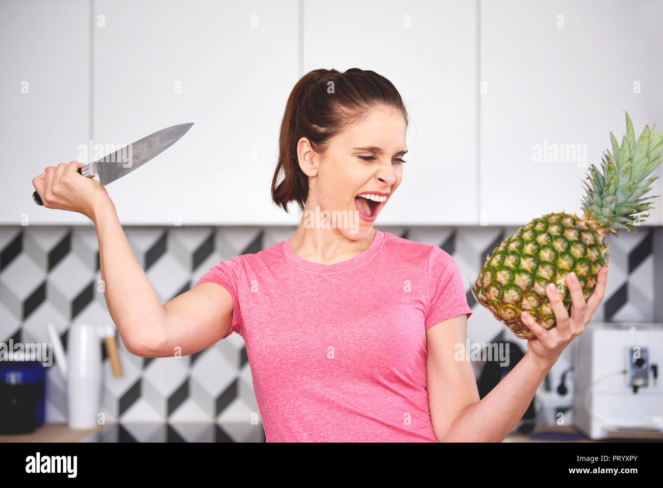 Portrait of screaming woman attacking pineapple with knife in the kitchen Stock Photo