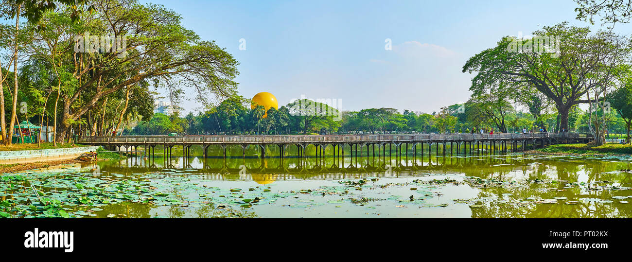 YANGON, MYANMAR - FEBRUARY 27, 2018: Panorama of Kandawgyi Park with old wooden bridge across the lake, covered with lotus plants and surrounded by sp Stock Photo