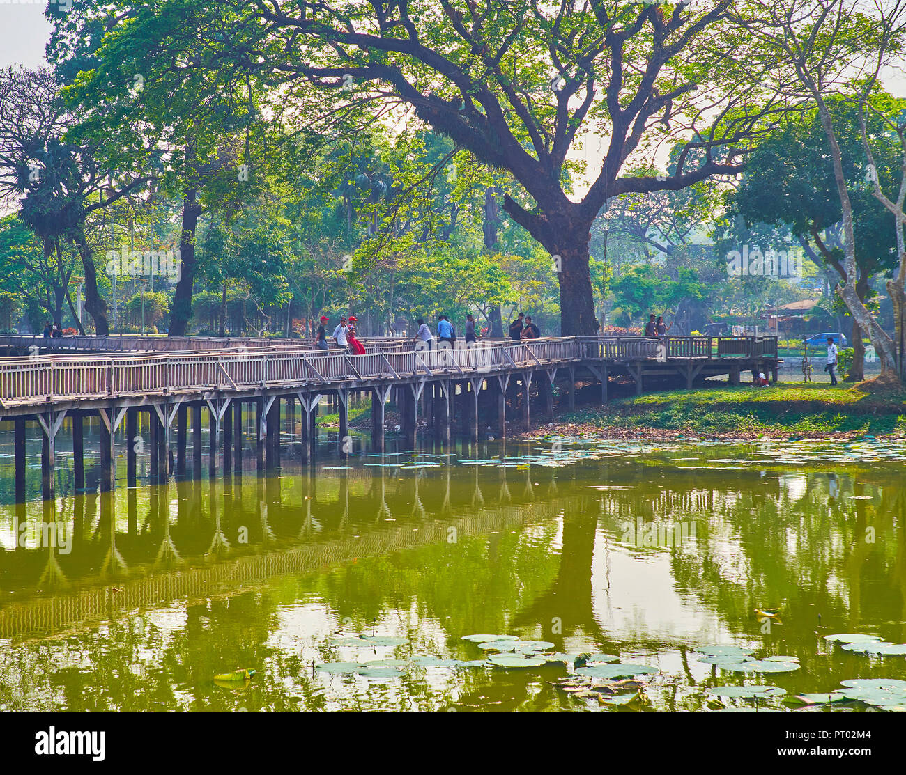 YANGON, MYANMAR - FEBRUARY 27, 2018: The pleasant daily walk along embankment of Kandawgyi Lake with a view on the old timber bridge and sprawling hta Stock Photo