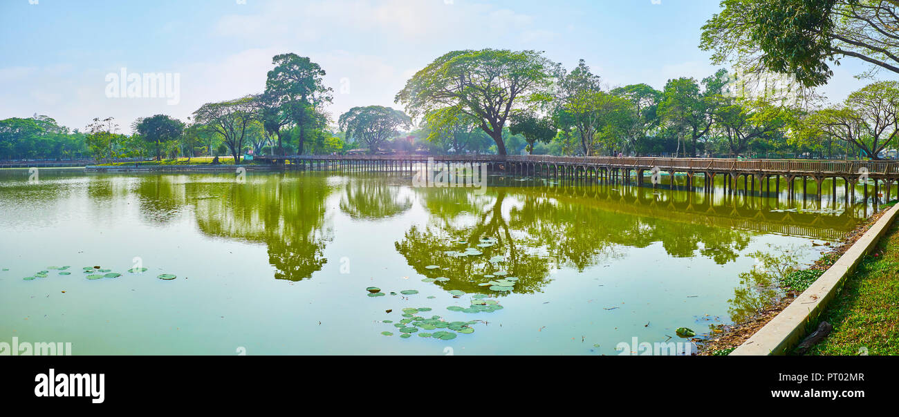 Panorama of Kandawgyi Lake with old wooden bridge, sprawling trees and blue sky, reflected in waters of the lake, Yangon, Myanmar. Stock Photo