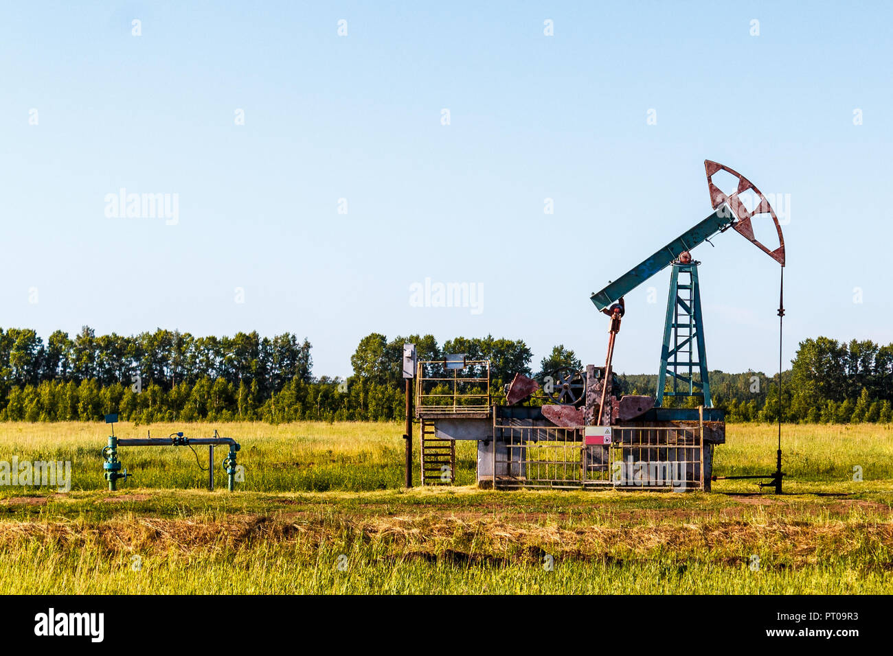 The oil pump works in summer in a green field against a background of green trees and a blue sky. Stock Photo