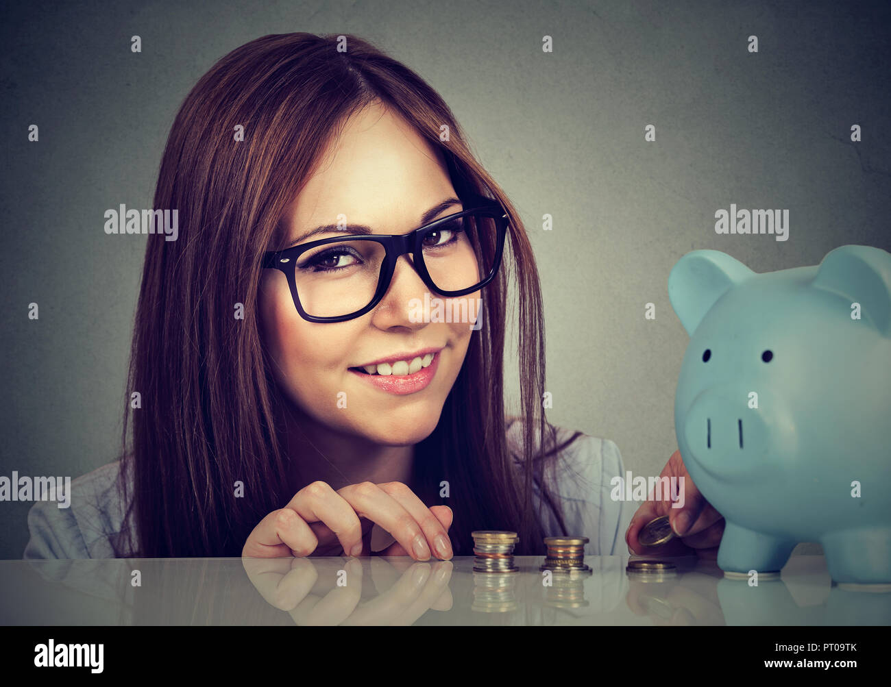 Young brunette in glasses at desk with blue piggy bank saving money looking at camera Stock Photo