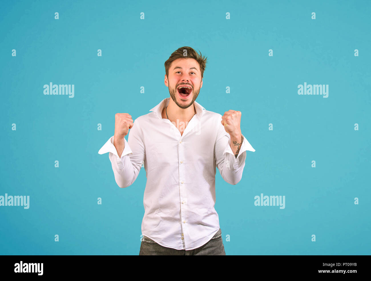 Excited handsome man in white shirt holding fists up and screaming with happiness on blue background Stock Photo