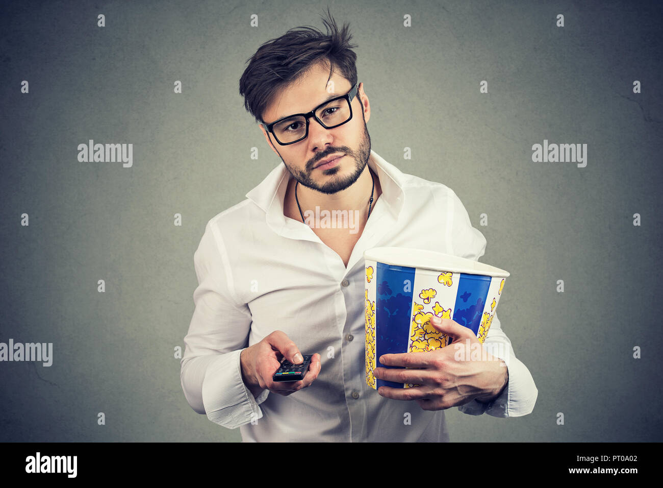Young handsome man in glasses looking bored while choosing TV show and holding bucket of popcorn Stock Photo
