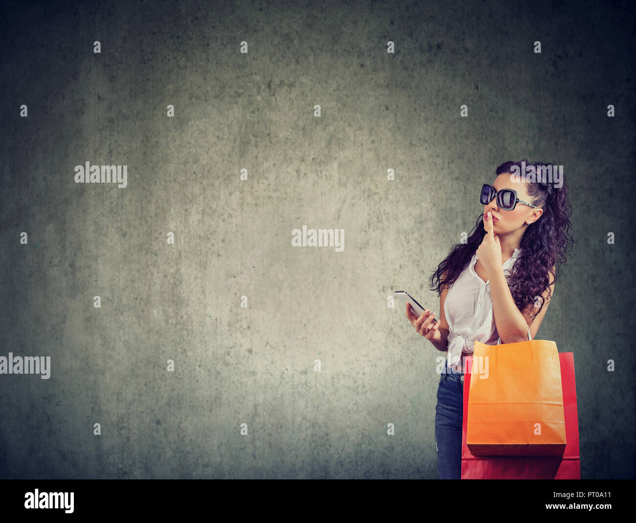 Trendy woman in sunglasses holding paper bags with smartphone and thinking with finger on lips Stock Photo