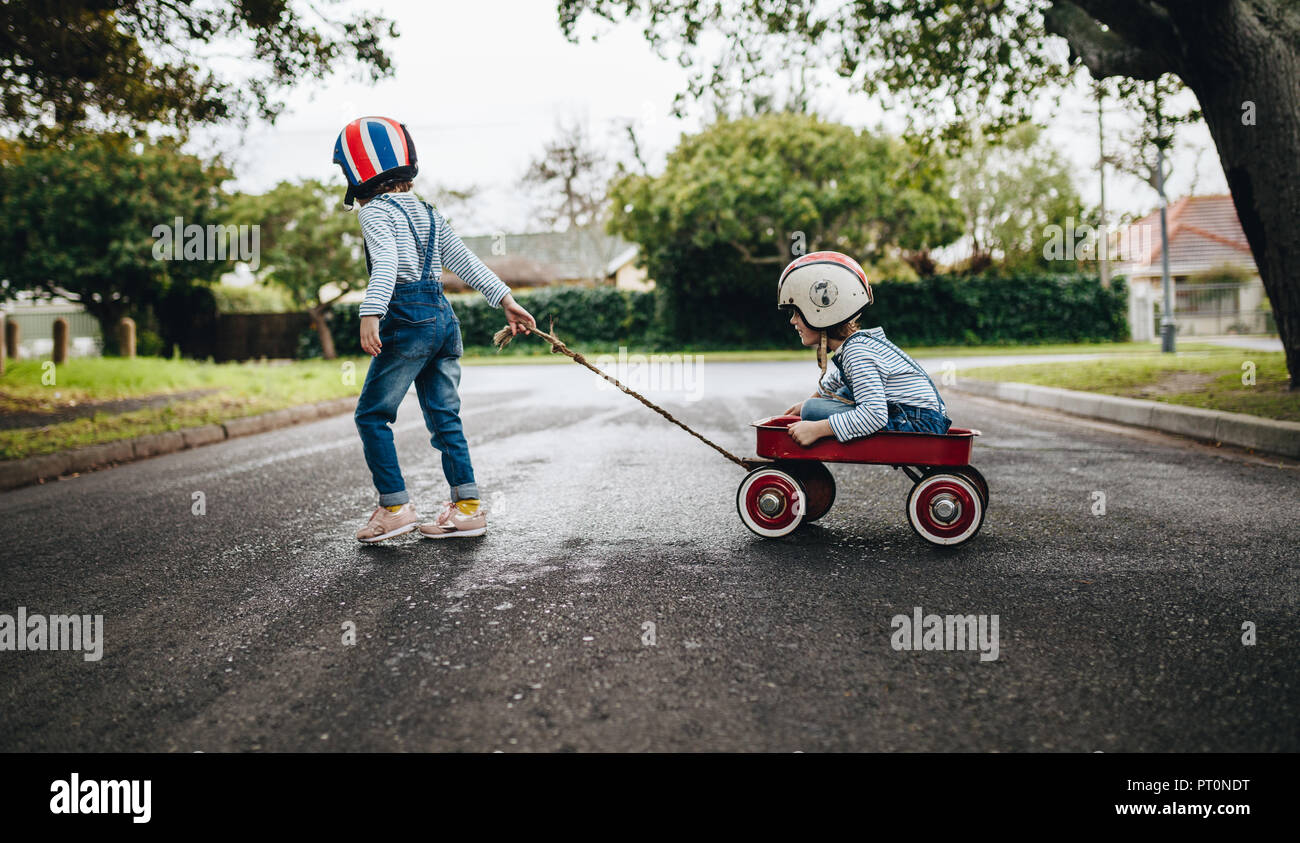 Little girl wearing helmet pulling her sister sitting in a wagon cart on the road. Kids playing outdoors with toy trolley. Stock Photo
