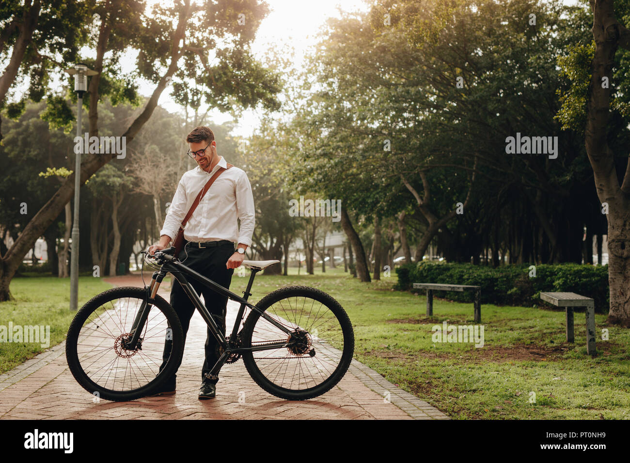 Man wearing office bag standing in park with his bicycle while commuting to office. Smiling businessman looking at his bike standing in the park. Stock Photo