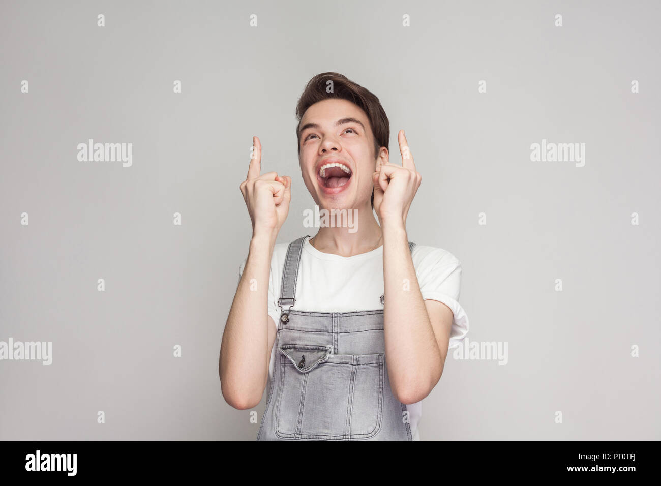 Portrait of happy amazed or excited young brunette man in casual style with t-shirt and denim overalls standing and looking and pointing in top of hea Stock Photo