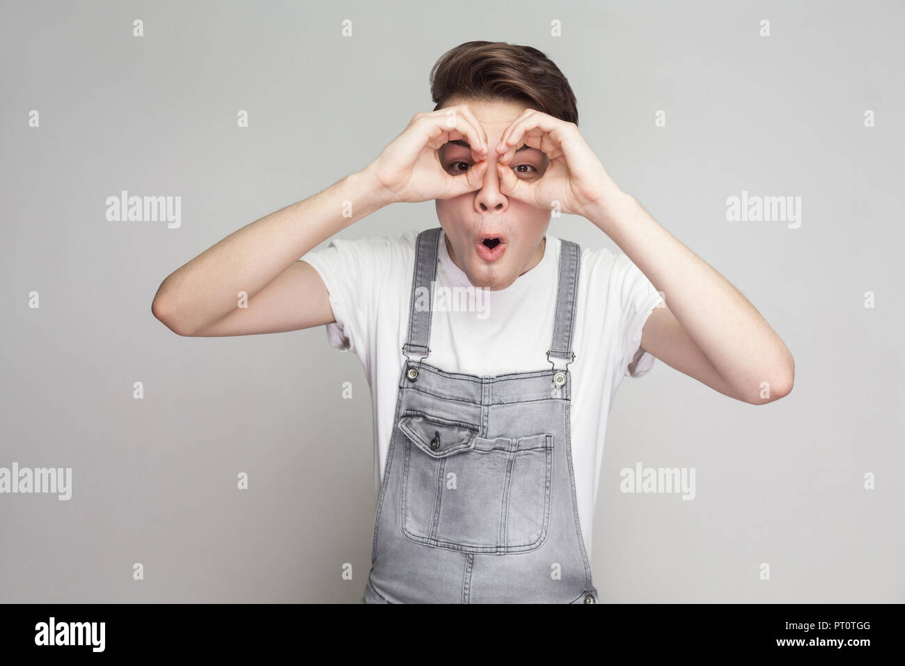 Portrait of amazed young brunette man in casual style with white t-shirt and denim overalls standing and looking at camera with binoculars gesture. in Stock Photo