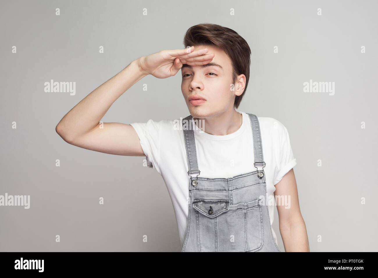 Serious brunette man in casual style with white t-shirt and denim overalls standing holding hands on forehead and looking to far with serious face. in Stock Photo