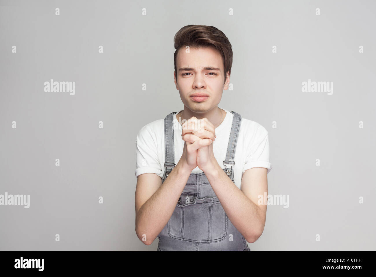 Portrait of worry hopeful young brunette man in casual style with denim overalls standing and looking at camera with pleading or forgiveness gesture.  Stock Photo