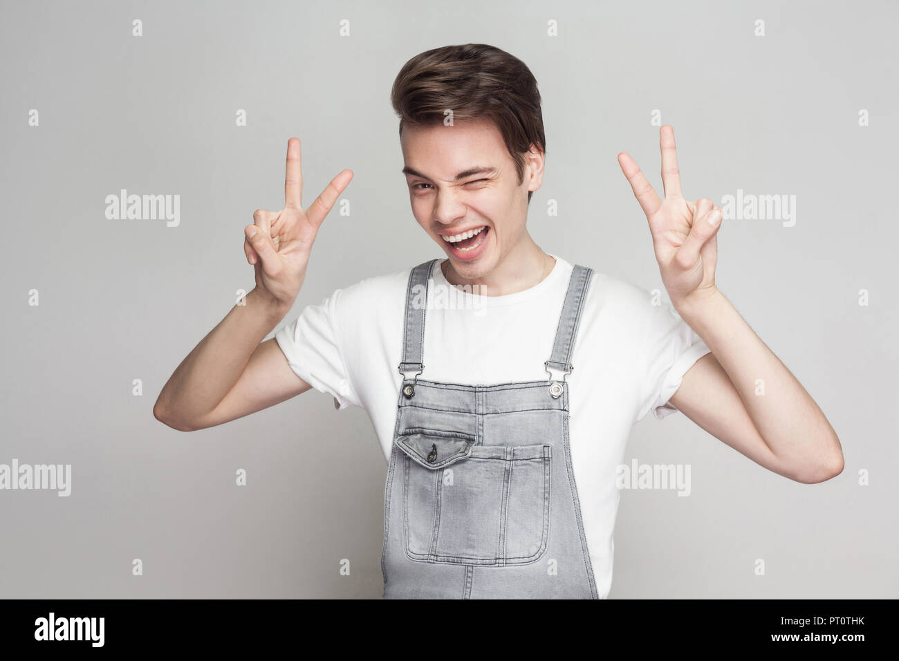 Portrait of funny young brunette man in casual style with denim overalls standing and looking at camera with victory or peace sign, winking and smilin Stock Photo