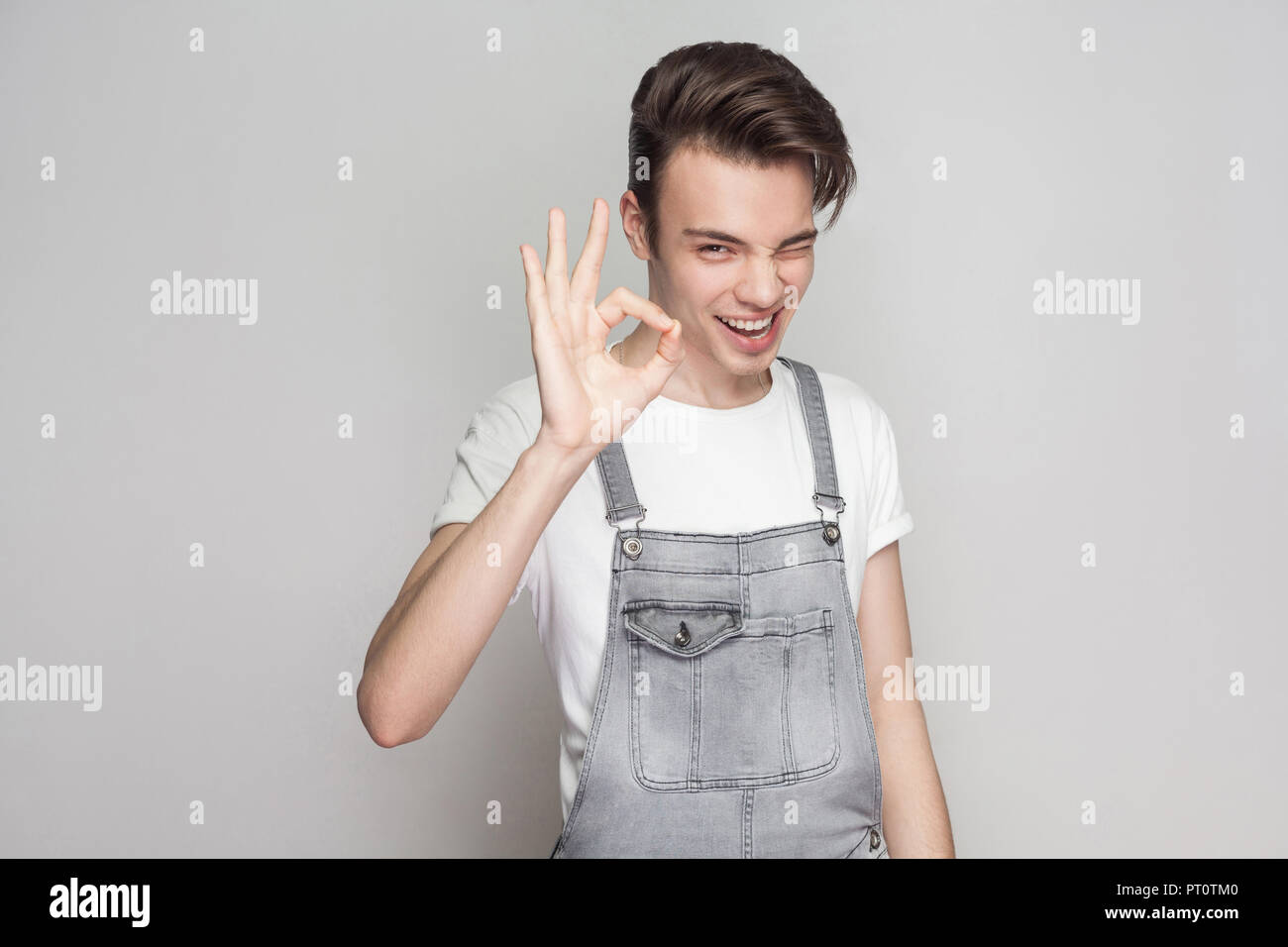 Happy young brunette man in casual style with t-shirt and denim overalls standing and looking at camera with toothy smile, winking and showing Ok sign Stock Photo