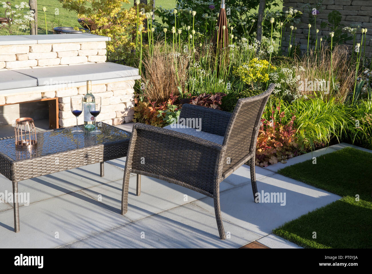 Small urban garden with stone patio with wicker garden patio furniture, coffee table and chair, stone bench, alliums, grasses, spring England UK Stock Photo