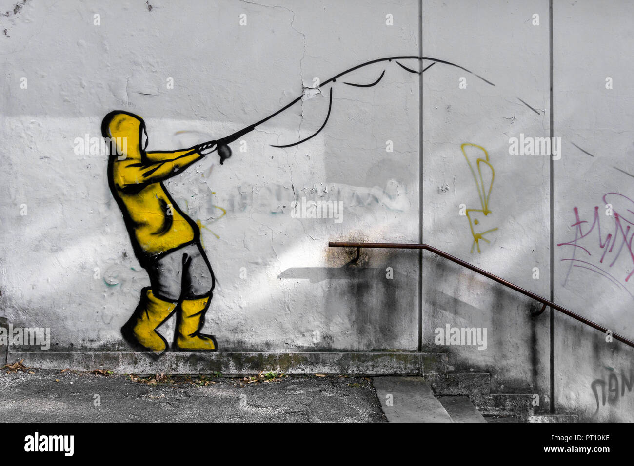 Street art by unknown artist on a concrete wall. The painting represents a boy fishing, suit with raincoat and yellow galoshes. Stock Photo