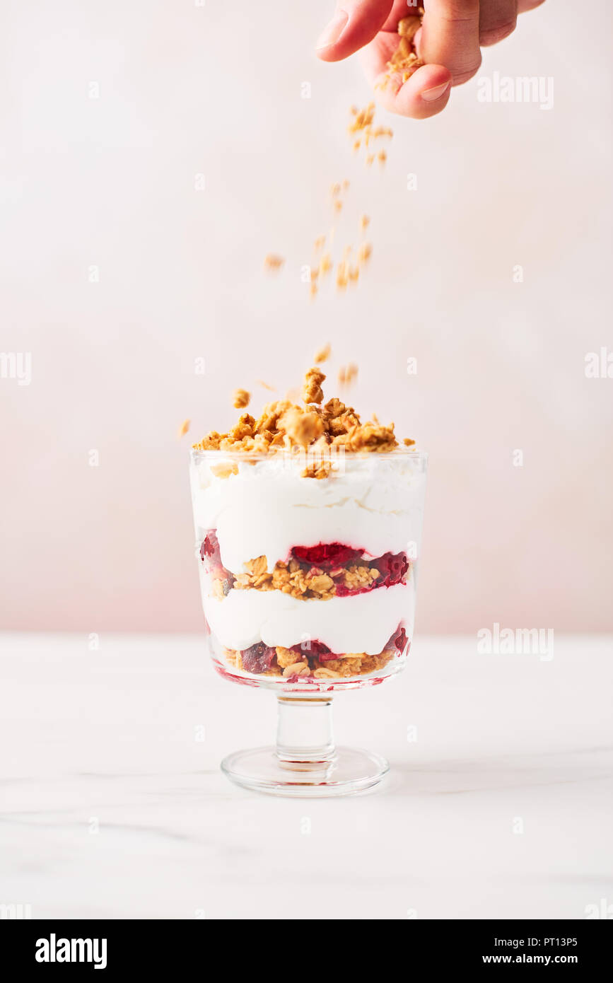 Hand sprinkling granola over healthy raspberry yogurt parfait in a glass on white marble table over pink background with copy space. Stock Photo