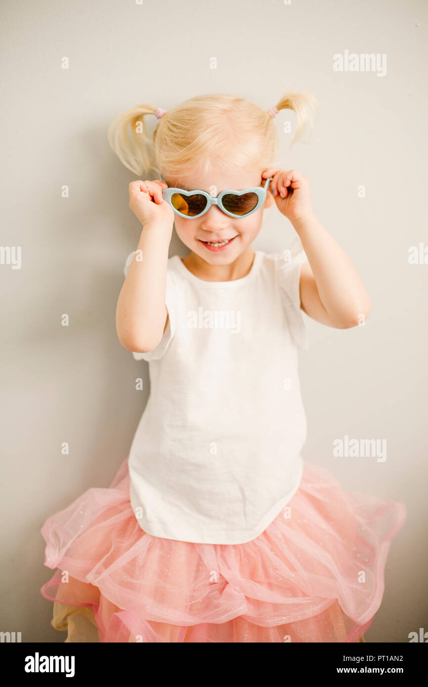 Portrait of blond little girl putting on heart-shaped sunglasses Stock Photo