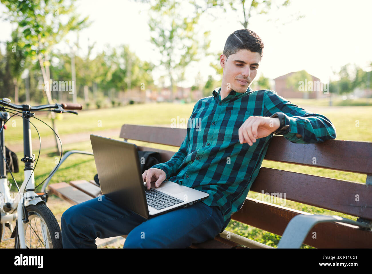 Young man with laptop on park bench checking the time Stock Photo