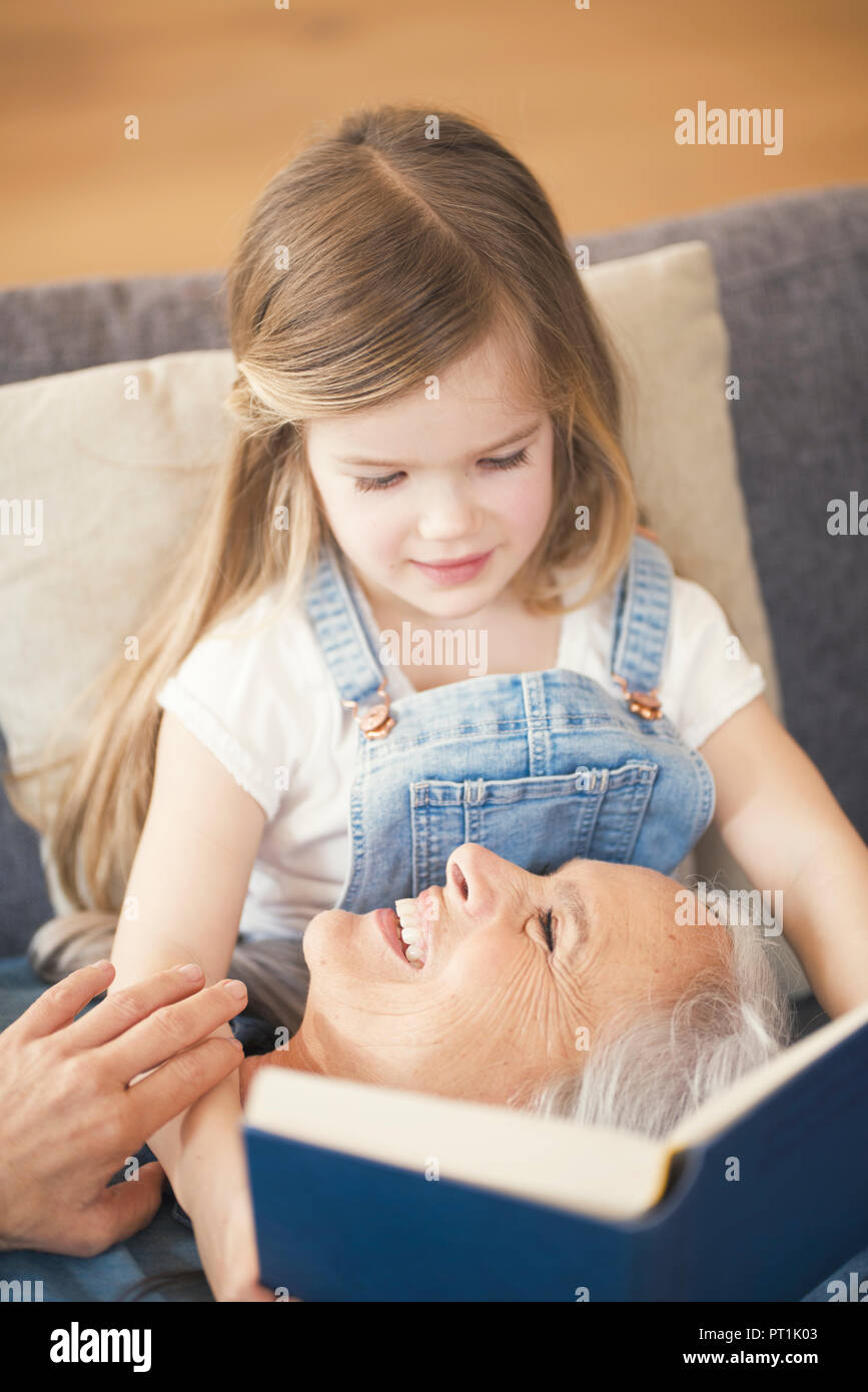 Grandmother and granddaughter sitting on couch, reading together a book Stock Photo