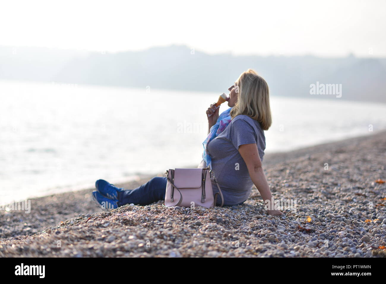A woman eating an ice cream and relaxing on the pebble beach during warm Indian summer weather at Seaton, Devon, England, UK Stock Photo