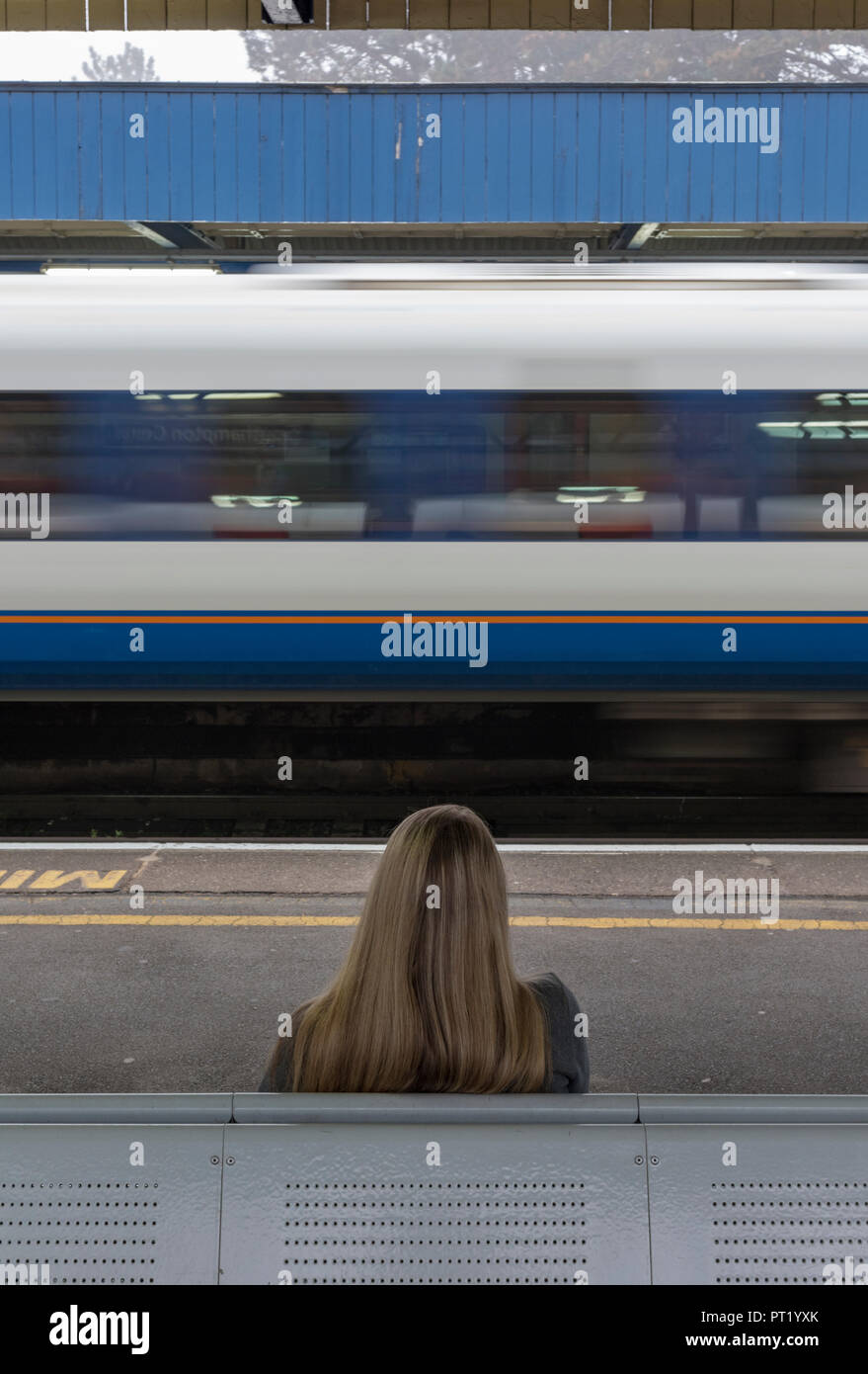 young woman sitting alone at a railway station with a train passing at speed, waiting for a train, the daily commute, young woman sitting on bench at railway station platform. Stock Photo
