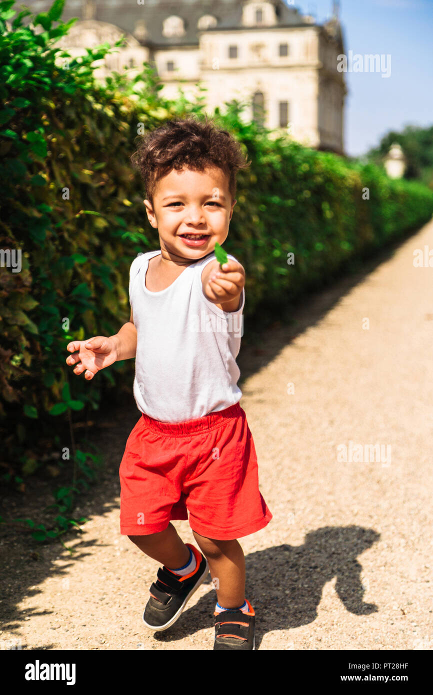 Cute little africanamerican or latinamerican boy in red shorts runnind with green leaf in park