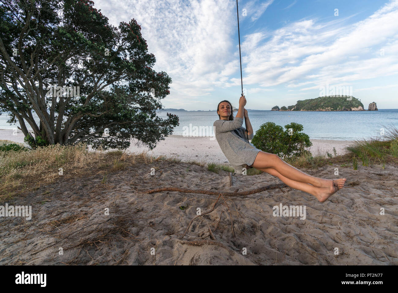 Young woman rope swinging on the beach and Te Karaka island in the background, Hahei, Waikato region, North Island, New Zealand, Stock Photo