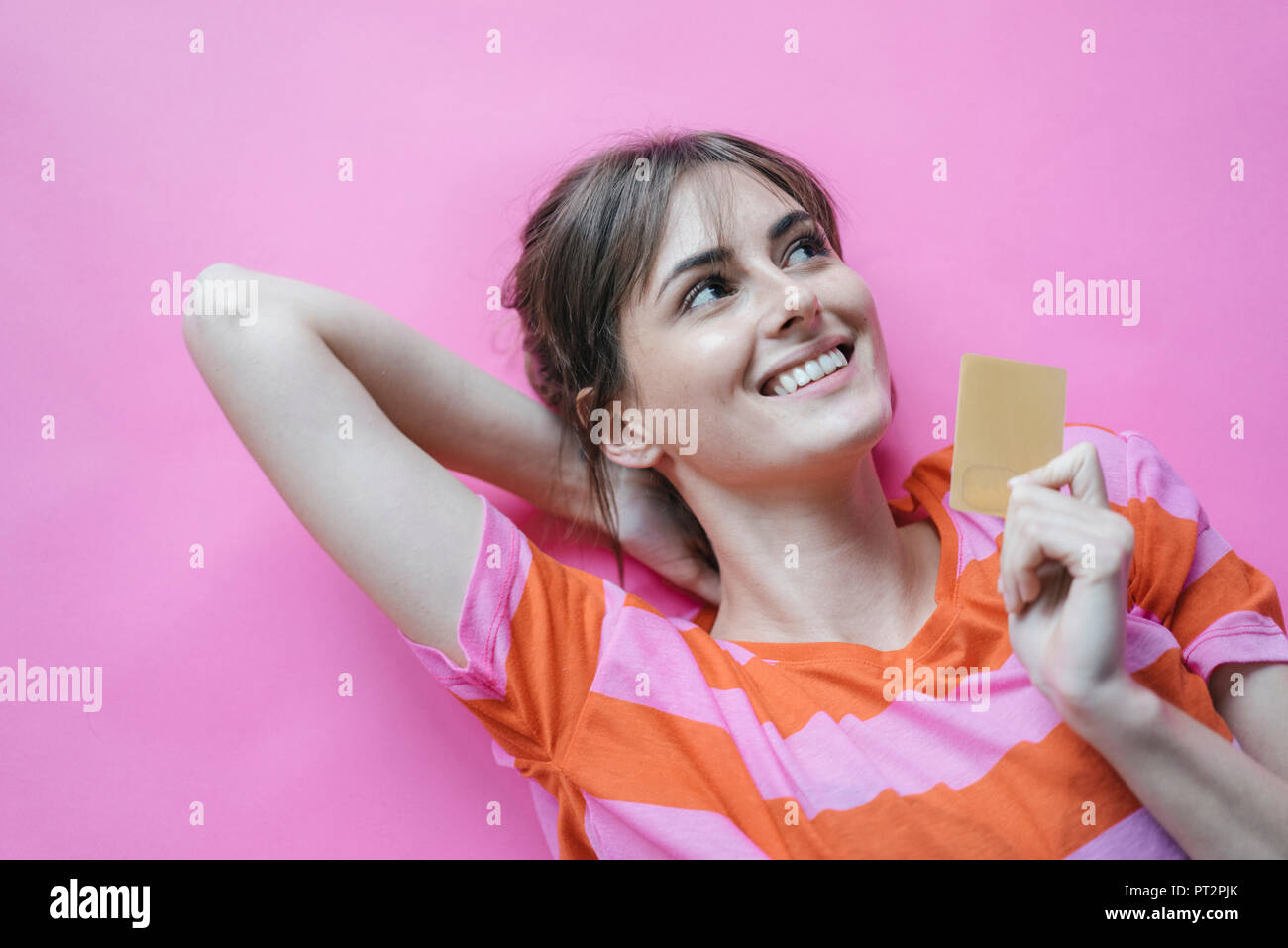 Woman lying on pink background with hands behind head, holding credit card Stock Photo