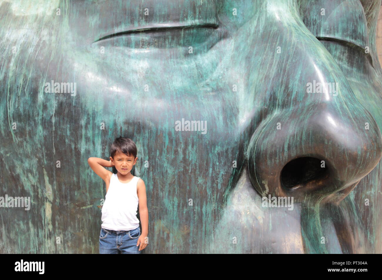 niño posando para la foto con una mano tocándose la cabeza recargado en una escultura verde de una cabeza a gran escala a plana luz del dia Stock Photo