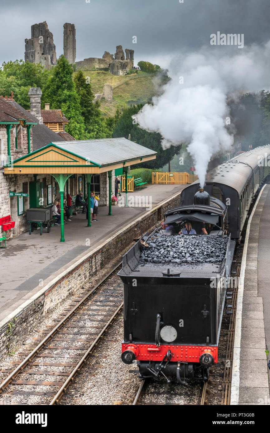 As threatening storm clouds gather above the castle, a steam locomotive pulls into the picturesque station at Corfe in Dorset. Stock Photo