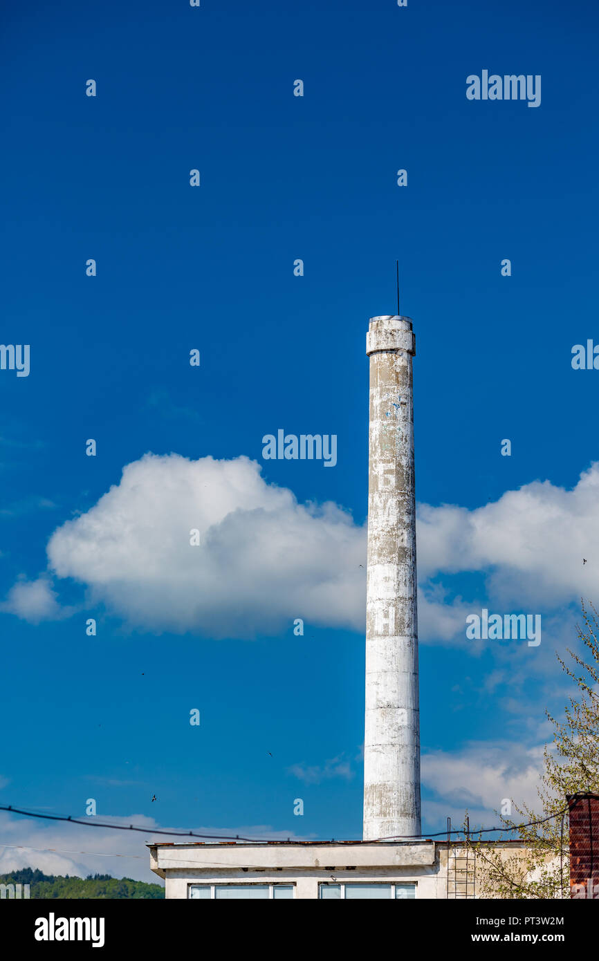 White painted smokeless concrete chimney with lightening rod of an abandoned Bulgarian factory against blue sky in a sunny spring day with white fluffy clouds Stock Photo