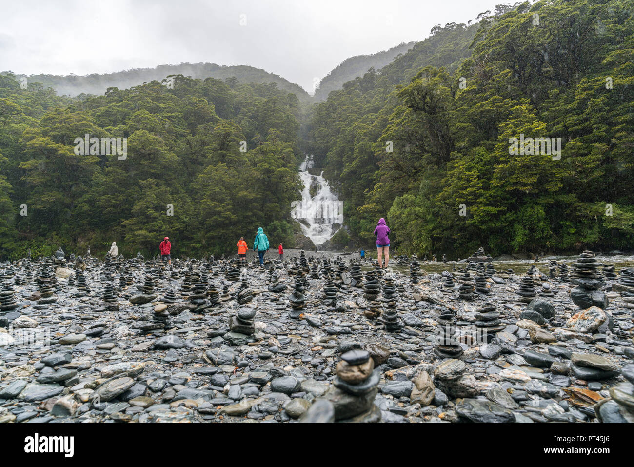Tourists walking through rock cairns at Fantail Falls on a rainy day, Mount Aspiring National Park, West Coast region, South Island, New Zealand, Stock Photo