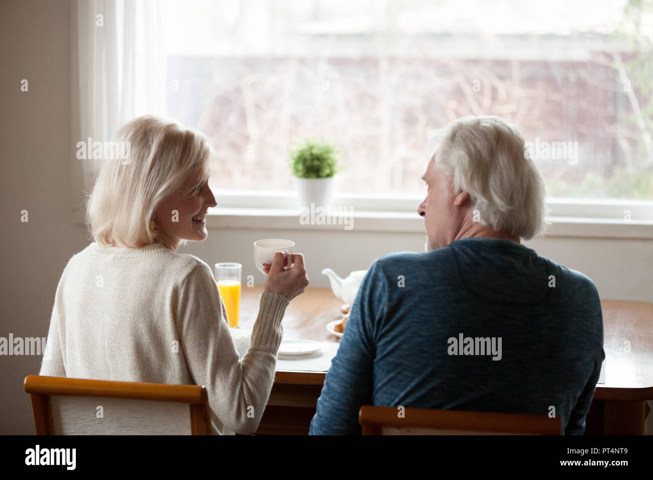 Rear view at happy senior old couple sitting at dining table drinking tea and talking at home, smiling middle aged mature family enjoying coffee and p Stock Photo