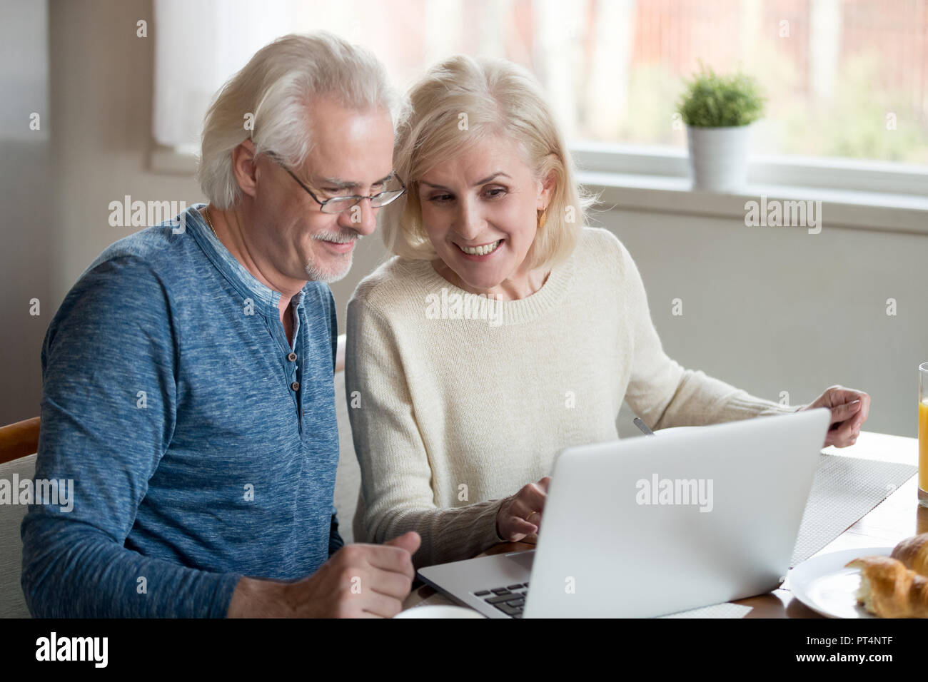 Happy old family couple talking using laptop having breakfast together, surprised excited senior woman looking at computer screen showing smiling midd Stock Photo