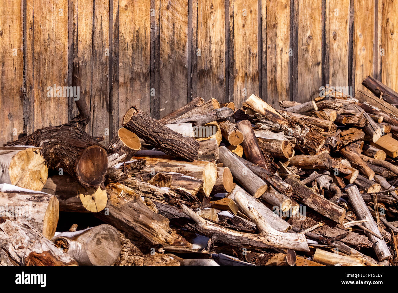 Large pile of various types of cut wood branches and logs, some with bark some not, is stacked against the wall of a building. The building has been w Stock Photo