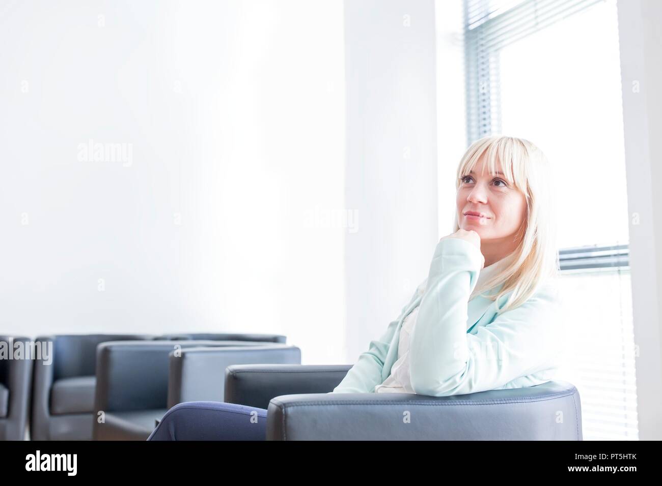 Pensive mid adult woman sitting in doctor's waiting room with hand on chin. Stock Photo