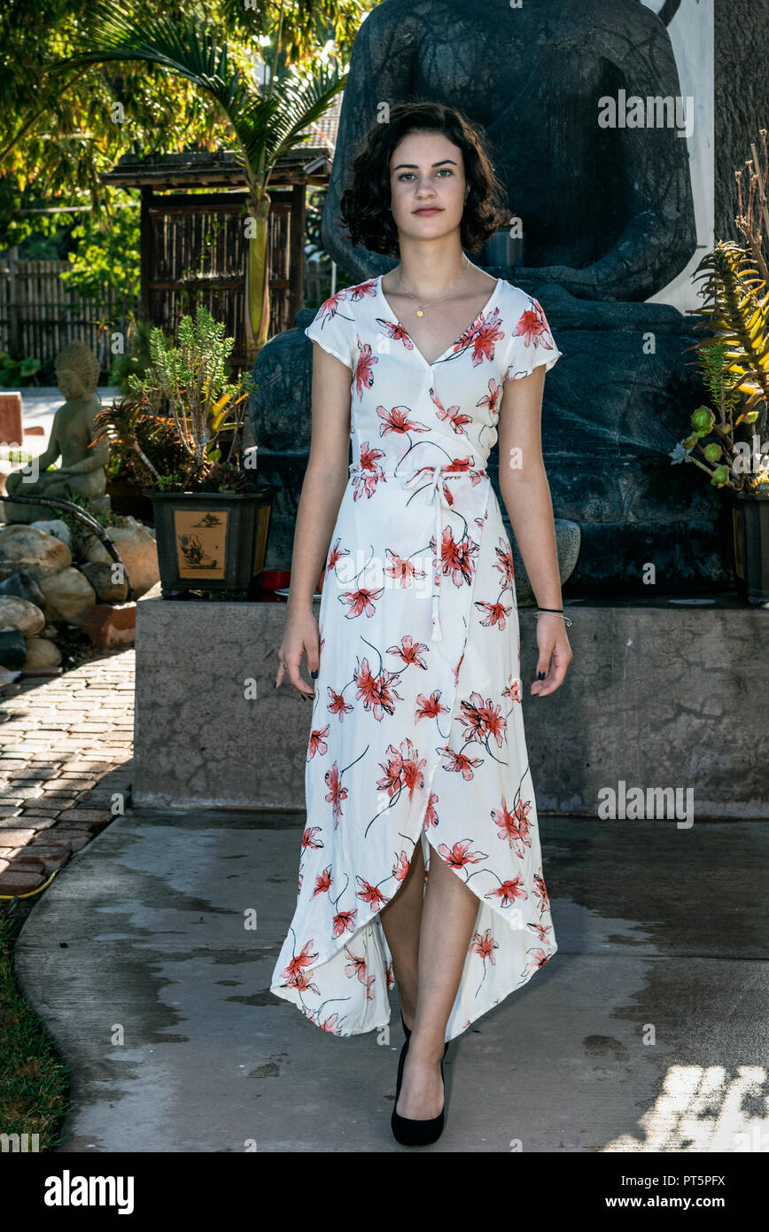 Beautiful young teen dressed in long flower print dress walking along concrete pathl in garden. Stock Photo