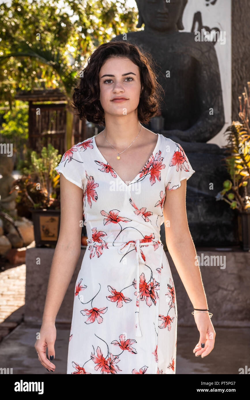 Headshot of beautiful young teen dressed in long flower print dress walking along concrete pathl in garden. Stock Photo