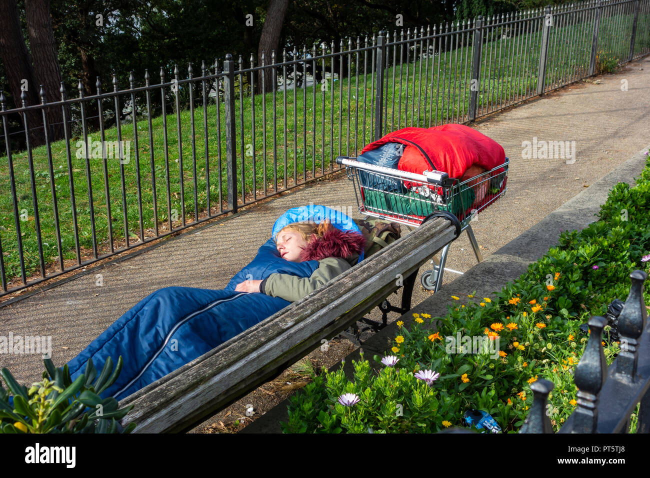 Early morning young homeless woman sleeping rough on a park bench, her only possessions in a shopping trolley. Stock Photo
