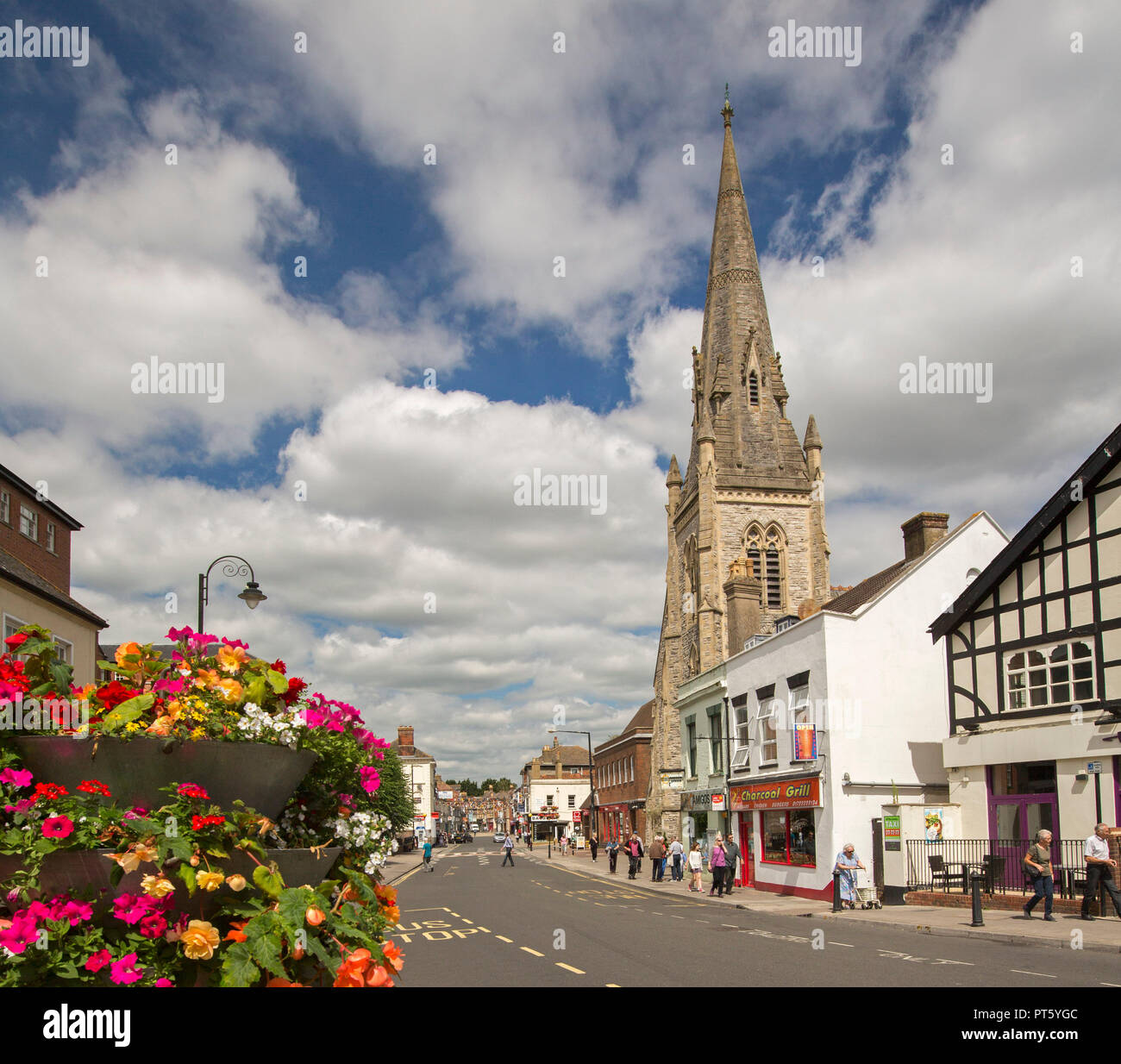 Main street in historic English town of Salisbury with shops, people strolling along footpath, colourful flowers & church spire reaching into blue sky Stock Photo