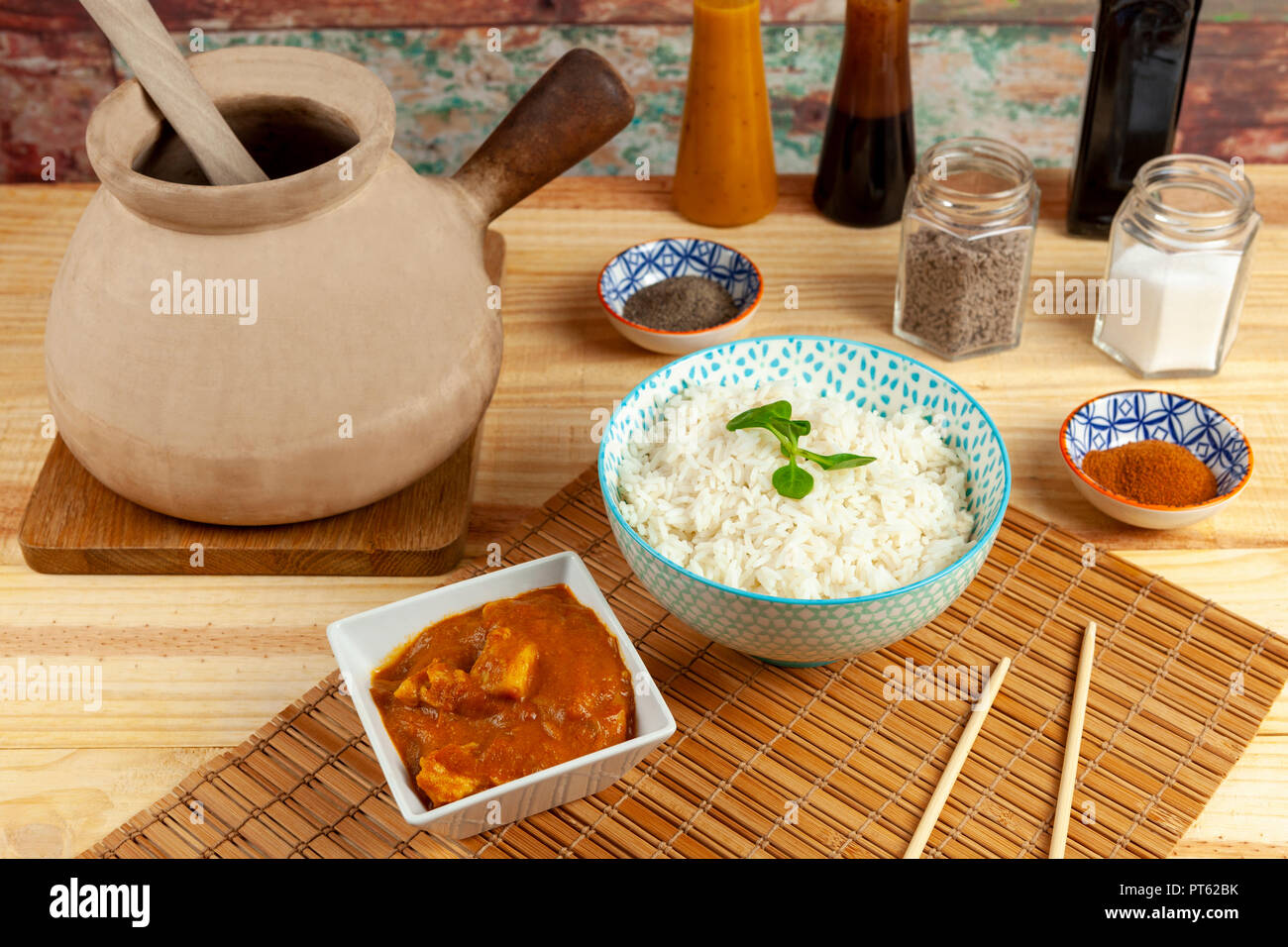 Bowl of rice and a portion of chicken curry laid out on a bamboo mat along side a traditional Chinese cooking pot and condiments on a wooden table Stock Photo