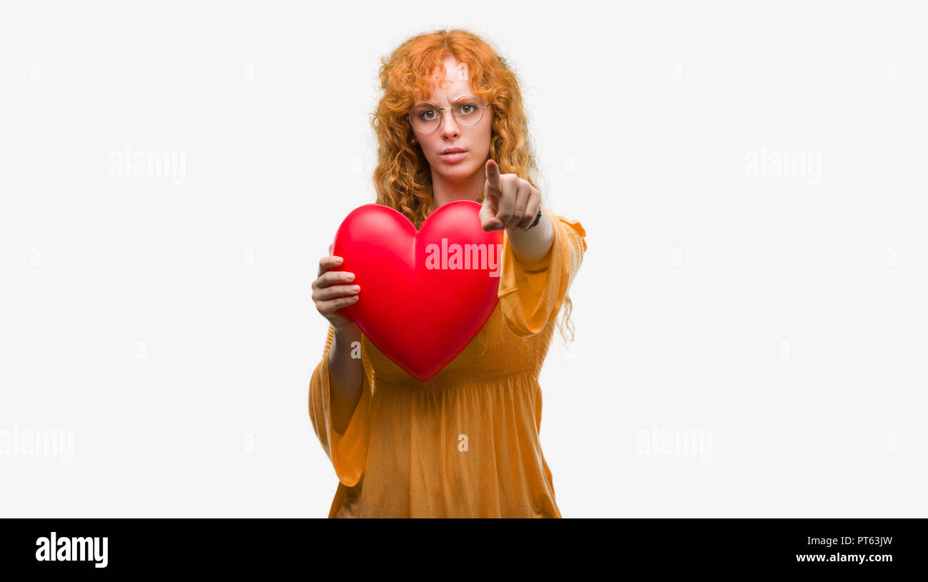 Young redhead woman in love holding red heart pointing with finger to ...