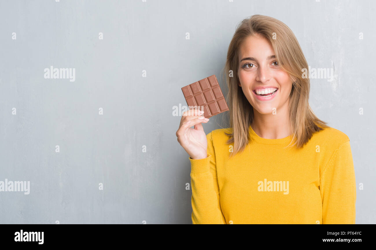 Beautiful young woman over grunge grey wall eating chocolate bar with a