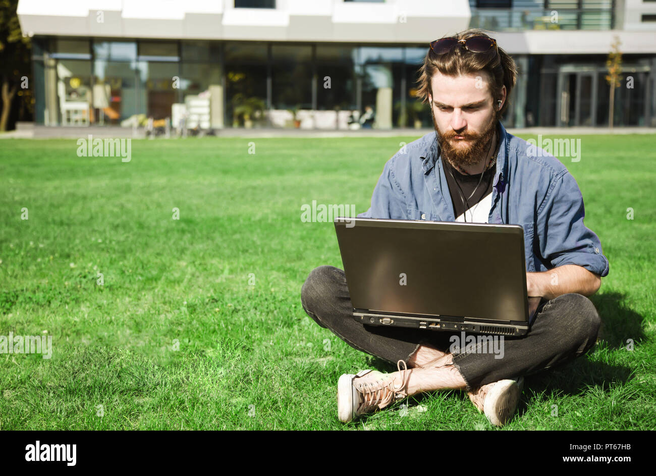 Handsome bearded student in jeans shirt wearing sunglasses sitting on the green grass near the university and doing his homework with laptop Stock Photo