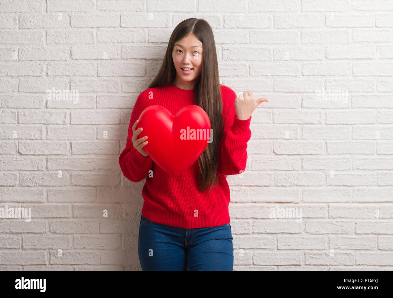 Young Chinese woman in love over brick wall holding red heart pointing ...