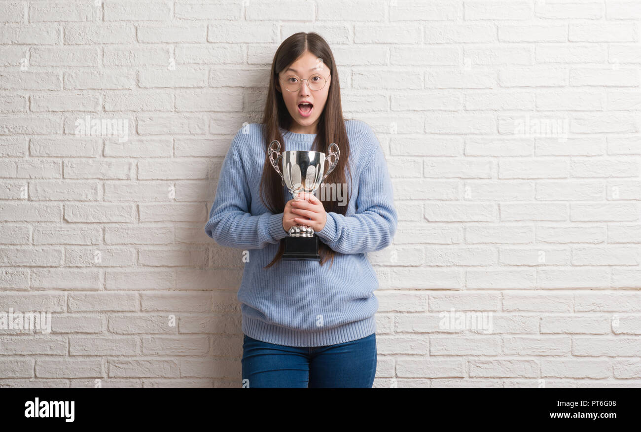 Young Chinese woman holding trophy scared in shock with a surprise face ...