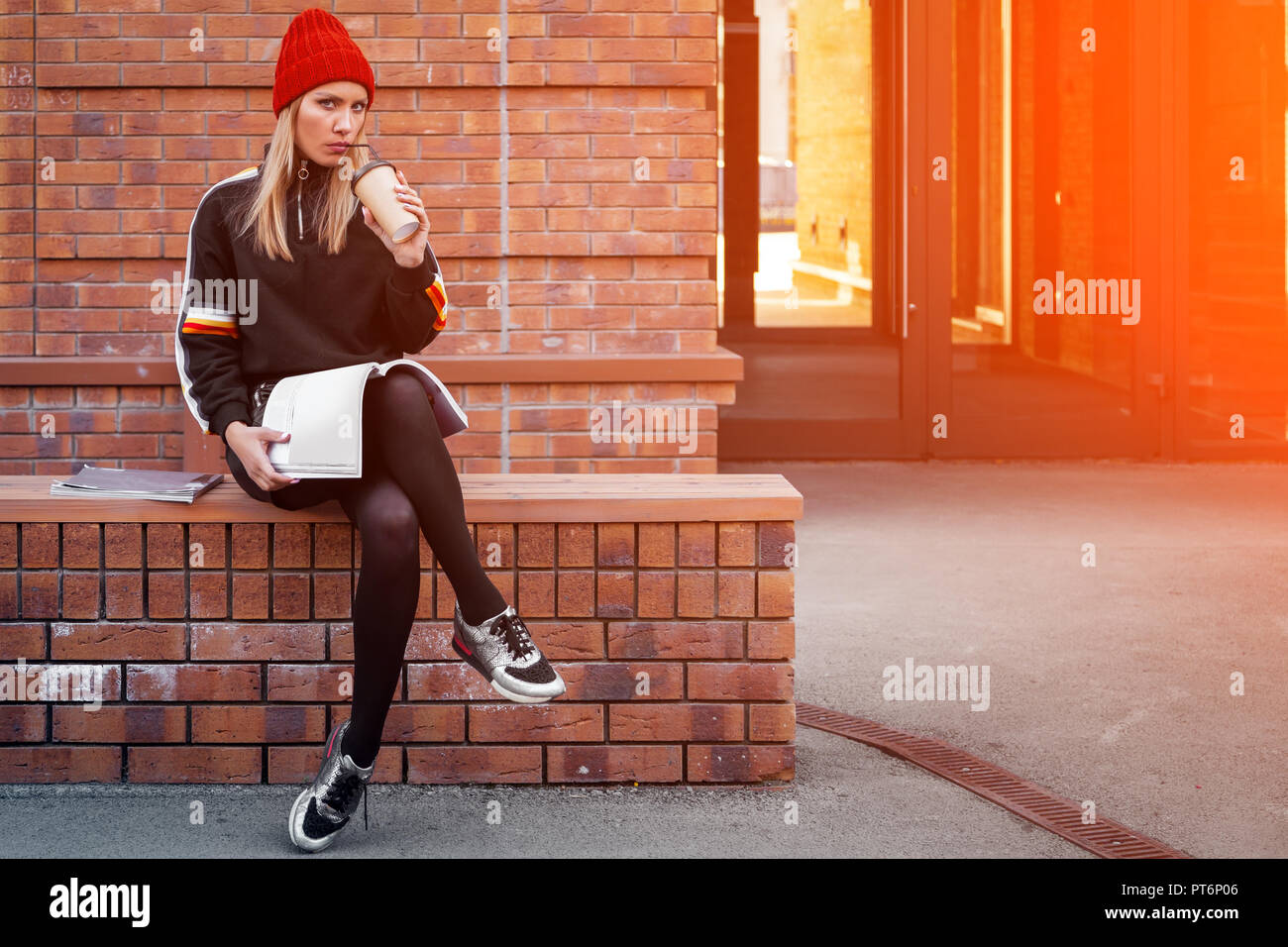 Fashion lifestyle portrait of young trendy woman dressed in black shirt,skirt and sneakers sits on bench, drink coffee to go, enjoy vacation  in the   Stock Photo