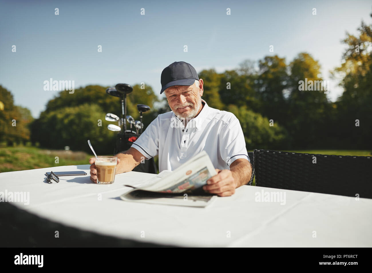 Smiling senior man sitting at a course restaurant drinking a coffee and reading a newspaper after playing a round of golf Stock Photo