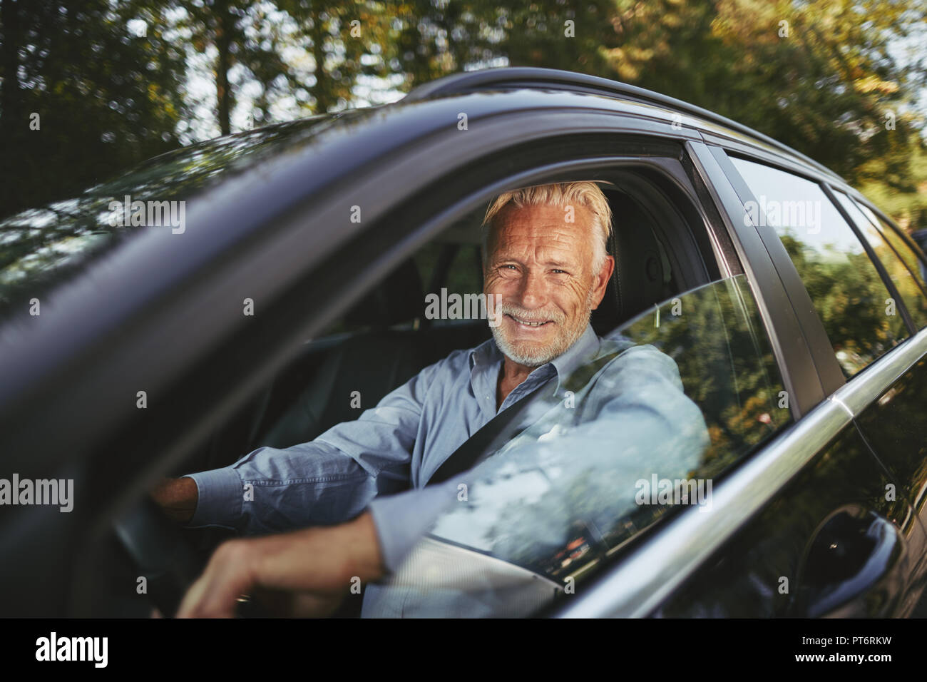 Senior man smiling while sitting in his car enjoying a drive along a tree lined road in the countryside Stock Photo