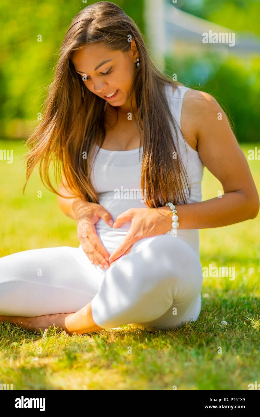 Beautiful pregnant woman sitting outdoor making hand heart gesture on belly Stock Photo